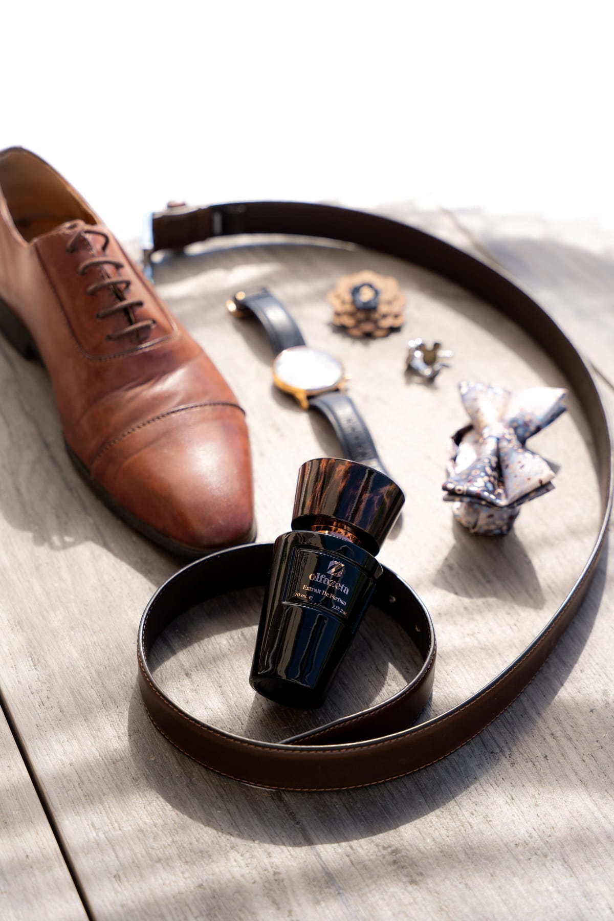 Flacon Olfazeta noir présenté avec une chaussure, une ceinture et une montre